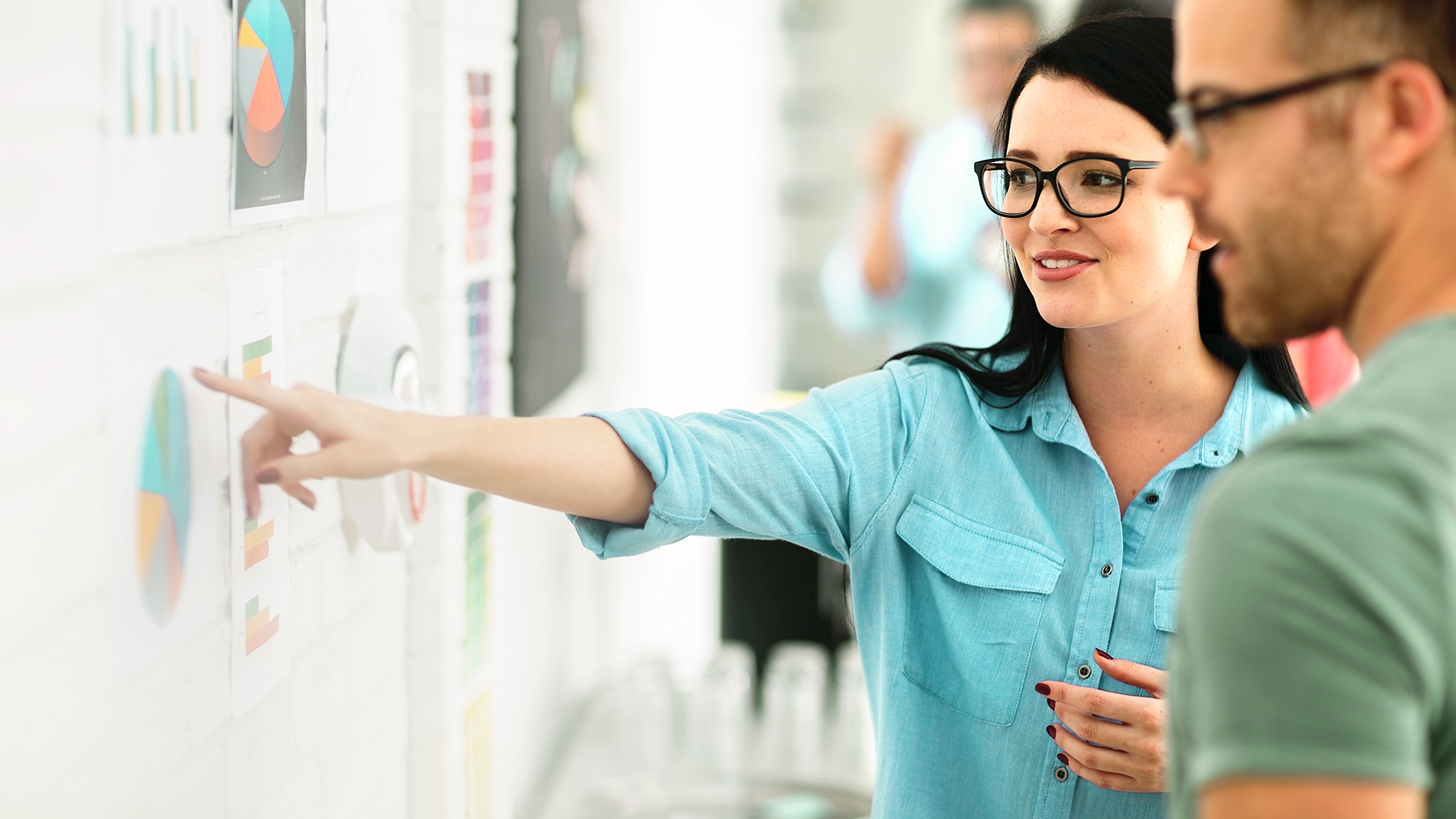 Eine Frau zeigt mit dem Finger auf eine Wand mit vielen Kuchendiagrammen. Ein Mann, unscharf im Vordergrund schaut zu.